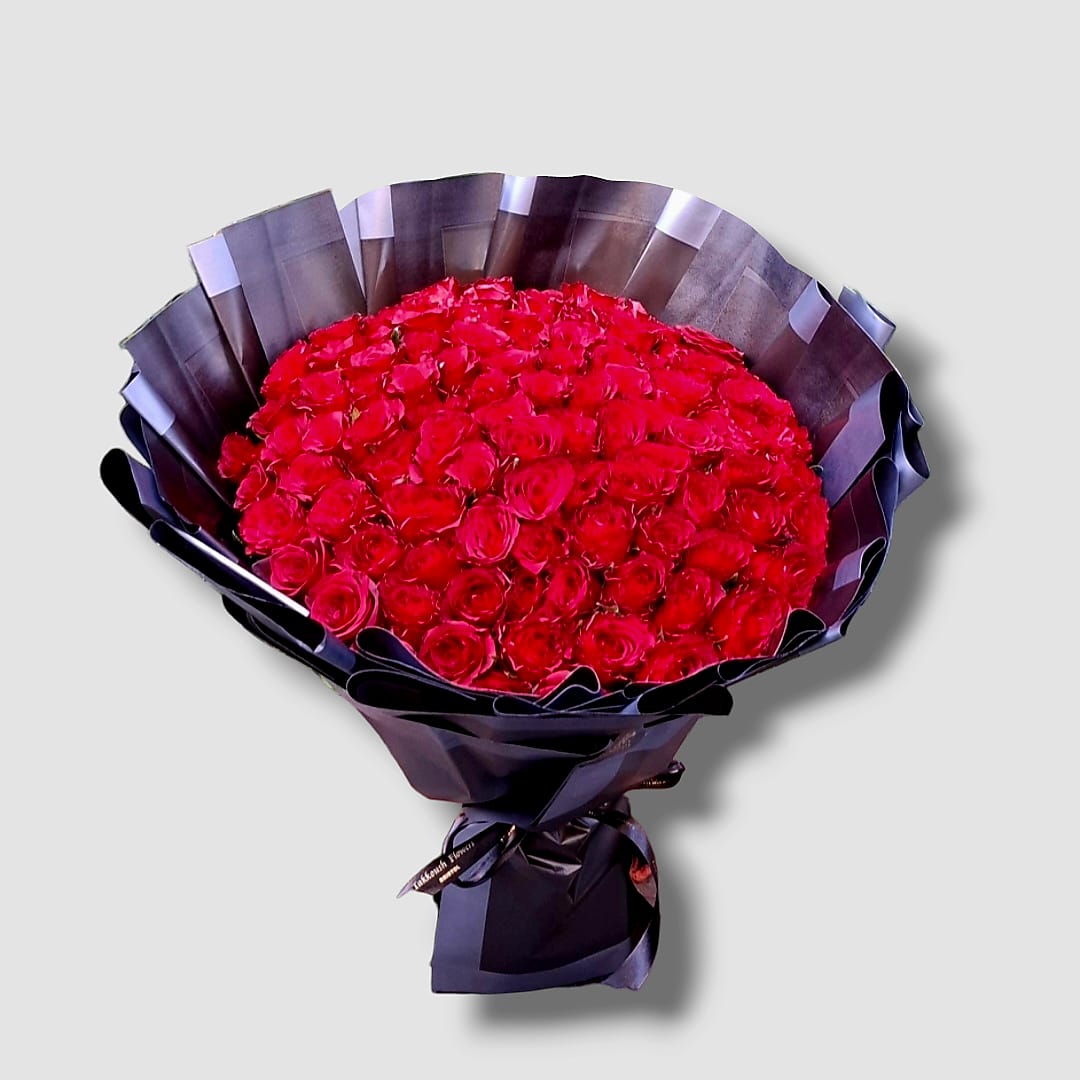 Opulent bouquet featuring 100 deep red long-stemmed roses densely arranged and wrapped in elegant matte black floral paper, tied with a matching black satin ribbon.
