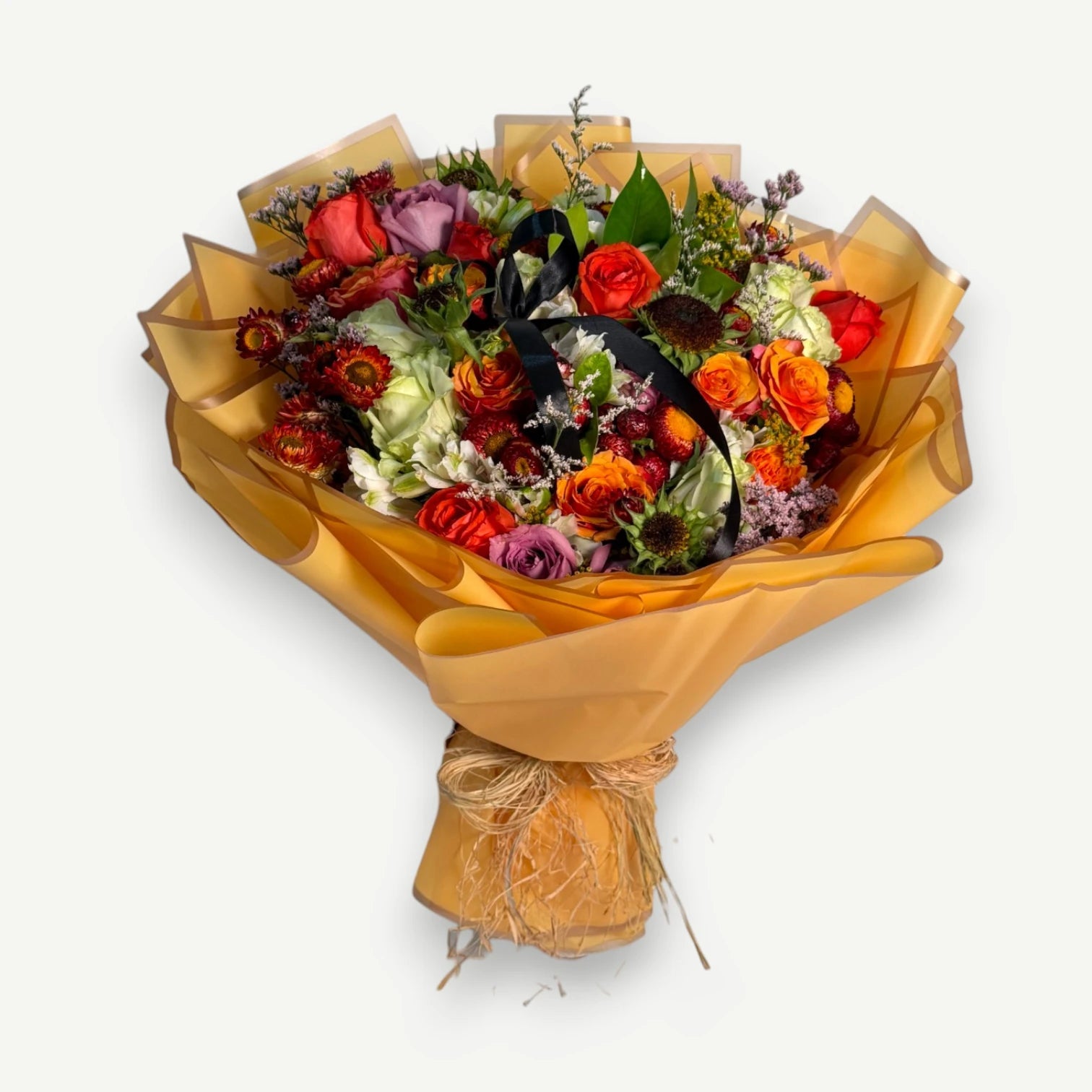 A vibrant mixed flower bouquet wrapped in golden-yellow paper, featuring red, orange, pink, and purple flowers with green foliage, tied with a black ribbon and jute twine, set against a plain white background.