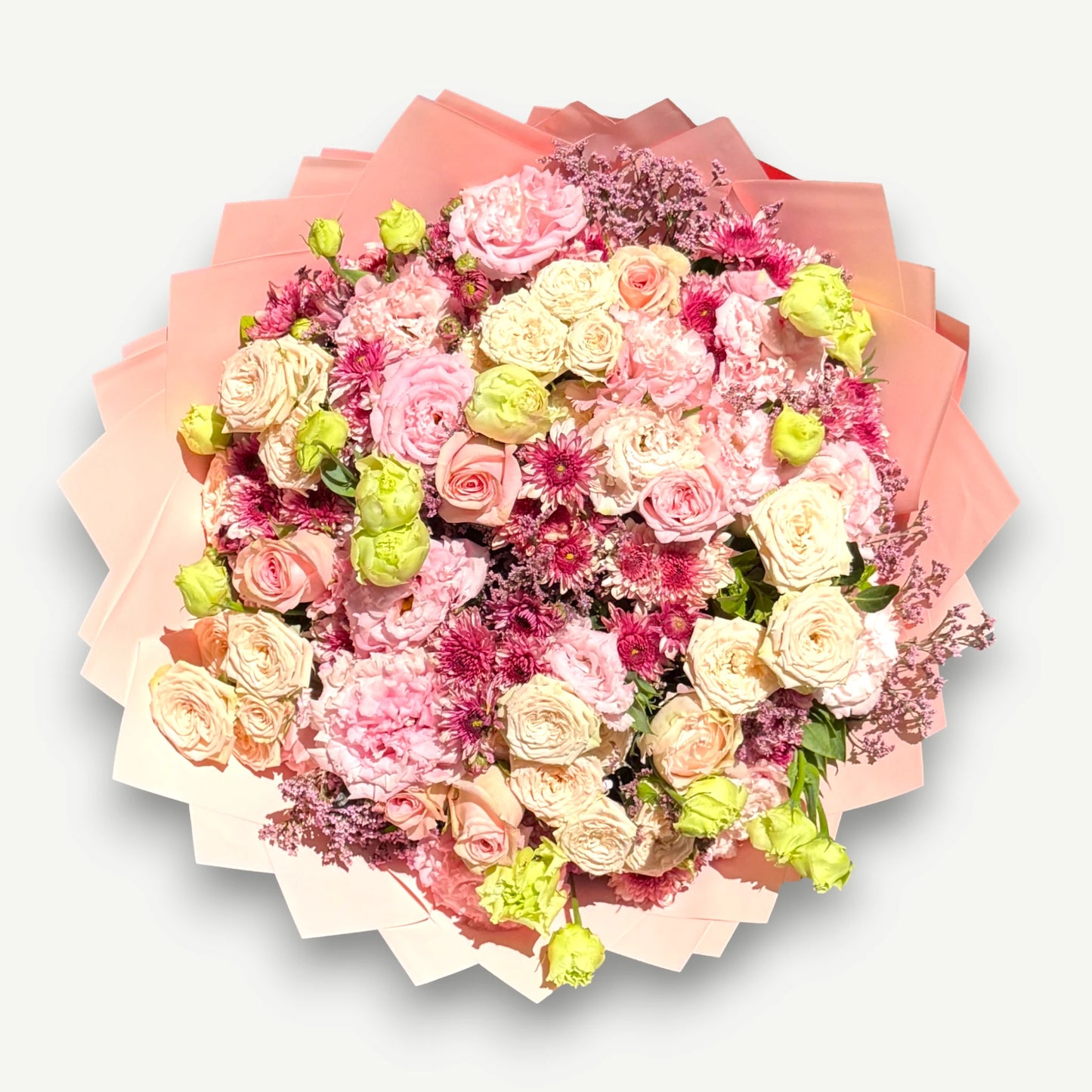 A large, round floral bouquet featuring pink, peach, cream, and green flowers, wrapped in layered pink and peach paper and photographed from directly above on a light gray background.