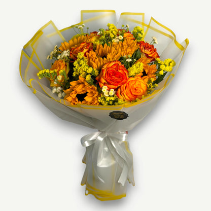 A vibrant floral bouquet wrapped in clear cellophane with yellow borders and tied with a white ribbon, set against a plain light gray background.