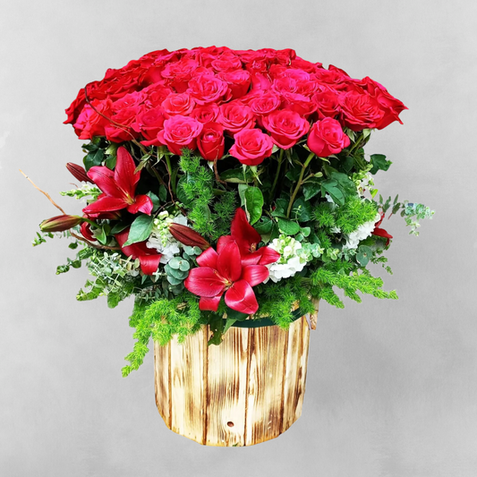 An image of a beautifully arranged bouquet of three dozens of lush, deep red roses nestled among three tall and elegant white lilies. The bouquet is displayed in a warm wooden box, adding a touch of natural beauty to the already stunning arrangement.